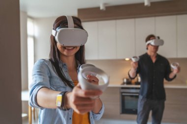 Genç, neşeli bir çift, sanal gerçeklik gözlükleriyle oynarken evlerinde kontrolörlerle oynuyorlar..