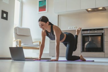 Genç ve güzel bir kız, evde, mutfağın arka planında spor yaparken laptopunu örnek egzersiz olarak kullanıyor..