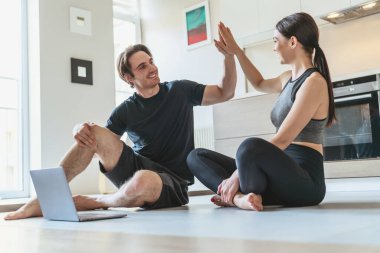 Genç bir çift, evlerinde mutfak arka planında spor yaparken ve örnek egzersizler için dizüstü bilgisayar kullanırken..
