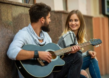 Man playing acoustic guitar while woman smiles and listens, sharing a relaxed moment together in an urban setting.