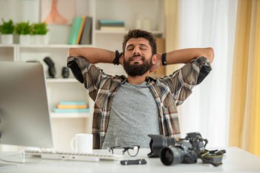 Sakallı adam kamera ekipmanıyla masada oturuyor, elleri başının arkasında arkana yaslanıyor, işten sonra dinlendiğini ve tatmin olduğunu söylüyor..