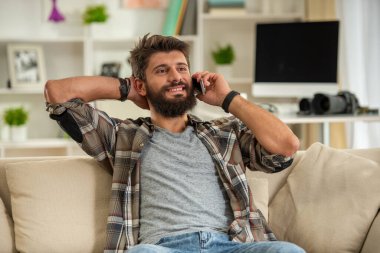 Kanepede oturan sakallı bir adam rahat bir duruşu, iletişimi ve rahat bir yaşam tarzını temsil eden akıllı telefonuyla konuşuyor..