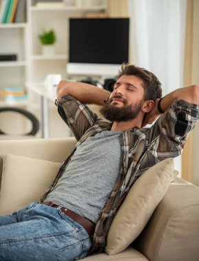 Sakallı genç adam gözleri kapalı bir kanepede uzanıyor, rahat bir evde dinleniyor, sakin huzurlu bir anın ve stres atmanın tadını çıkarıyor..