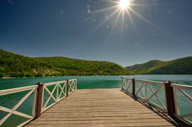 Yeşil tepeler ve turkuaz göl manzarası gün boyunca küçük bir rıhtımdan çekilmiş.