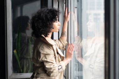 Genç güzel kız pencerenin yanında yağmurlukla duruyor.