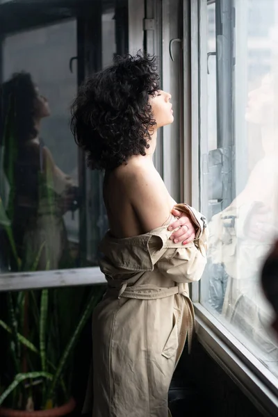young beautiful woman stands near the window in a raincoat