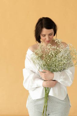 Stüdyoda poz veren genç ve güzel bir kadın sarı bir duvarın yanında beyaz çiçeklerle duruyor.