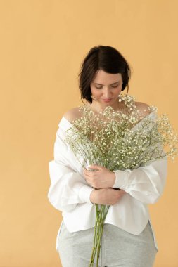 Stüdyoda poz veren genç ve güzel bir kadın sarı bir duvarın yanında beyaz çiçeklerle duruyor.