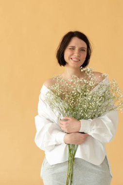 Stüdyoda poz veren genç ve güzel bir kadın sarı bir duvarın yanında beyaz çiçeklerle duruyor.