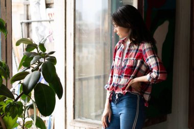 Stüdyoda poz veren genç, güzel bir kadın, ekose gömlek ve kot pantolonla pencerenin yanında duruyor.