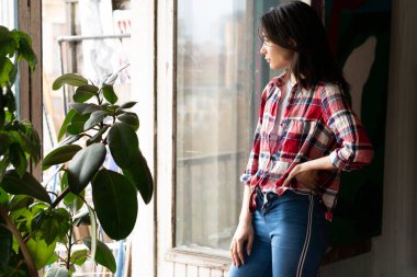 Stüdyoda poz veren genç, güzel bir kadın, ekose gömlek ve kot pantolonla pencerenin yanında duruyor.