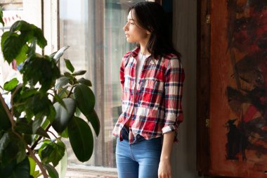 Stüdyoda poz veren genç, güzel bir kadın, ekose gömlek ve kot pantolonla pencerenin yanında duruyor.