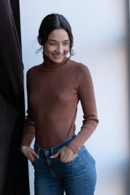 Stüdyoda poz veren genç, güzel bir kadın, pencerenin yanında duruyor.