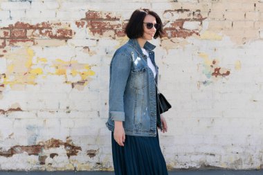 Dışarıda poz veren genç ve güzel bir kadın, şehirde kot ceketle dikiliyor.