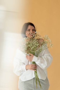 Stüdyoda beyaz çiçeklerle poz veren genç ve güzel bir kadın, sarı duvarın yanında duruyor.