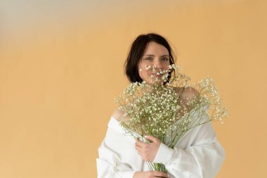 Stüdyoda beyaz çiçeklerle poz veren genç ve güzel bir kadın, sarı duvarın yanında duruyor.