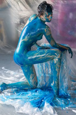 Genç ve güzel bir kadın çıplak poz veriyor, mavi boyayla boyanmış.