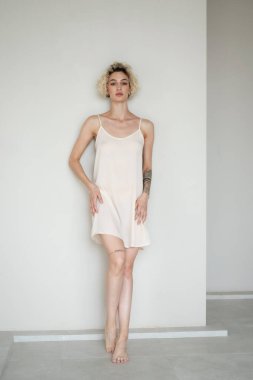 Stüdyoda beyaz bir gecelikle poz veren genç ve güzel bir kadın beyaz bir duvarın yanında duruyor.