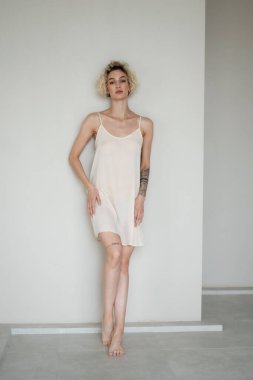 Stüdyoda beyaz bir gecelikle poz veren genç ve güzel bir kadın beyaz bir duvarın yanında duruyor.