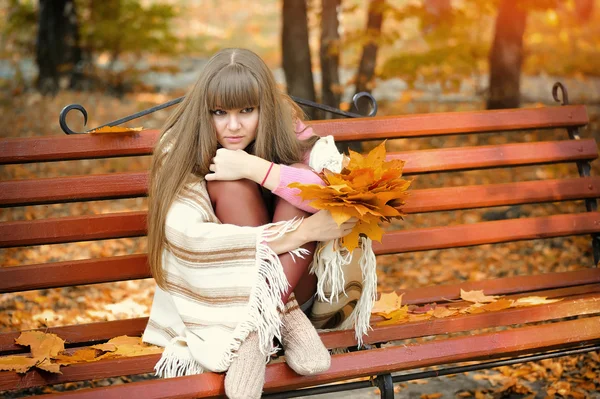 Schöne junge Frau sitzt auf einer Bank mit Herbstblättern — Stockfoto