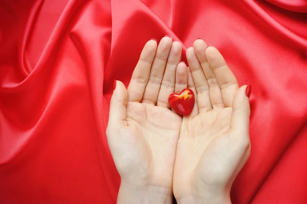 Coração decorativo nas mãos das mulheres contra um fundo de seda vermelha — Fotografia de Stock