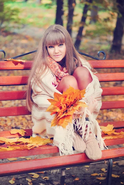 Schöne junge Frau sitzt auf einer Bank mit Herbstblättern — Stockfoto