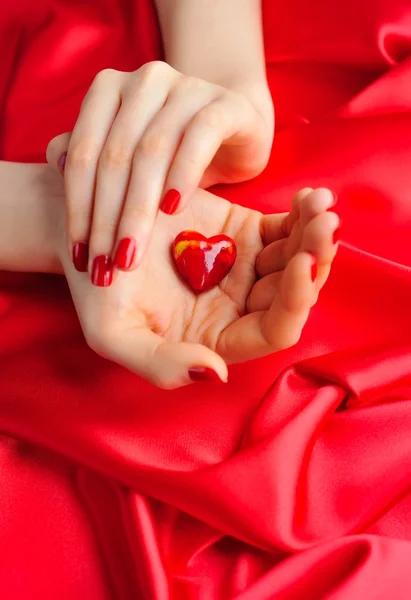 Cuore decorativo nelle mani delle donne su uno sfondo di seta rossa — Foto Stock