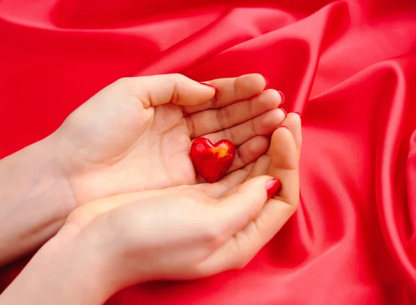 Coração decorativo nas mãos das mulheres contra um fundo de seda vermelha — Fotografia de Stock