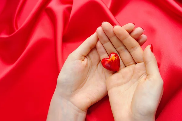 Coração decorativo nas mãos das mulheres contra um fundo de seda vermelha — Fotografia de Stock