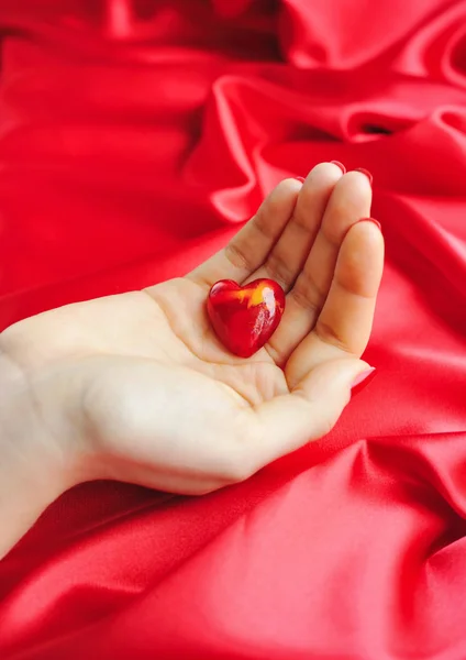 Coração decorativo nas mãos das mulheres contra um fundo de seda vermelha — Fotografia de Stock