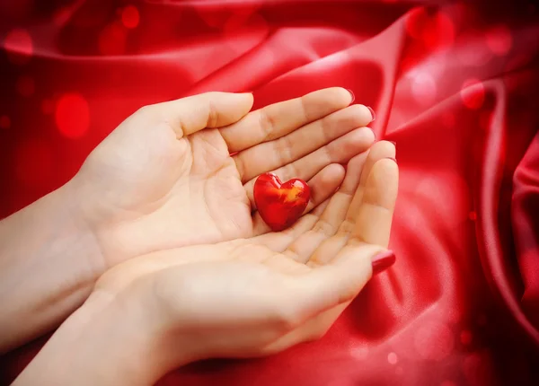 Corazón decorativo en las manos de las mujeres sobre un fondo de seda roja — Foto de Stock