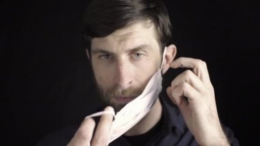 Siyah arka plandaki bir odada sakallı beyaz bir adam tıbbi bir maske takıyor ve düzeltiyor. Sağlık koruma konsepti. Viral hastalıkların yayılmasını önleme.