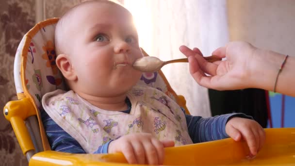 Mères main nourrissant bébé avec une cuillère à la chaise haute jaune tandis que petit garçon mignon regarde en arrière et puis regarder à la caméra 4K lent mo — Video