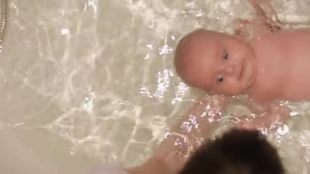 Schöne Neugeborene schwimmt lustig im Badezimmer mit Hilfe der Mutter. Schöner Gesichtsausdruck, dicker Hals, große Augen. Glückliche Kindheit, elterliche Fürsorge — Stockvideo