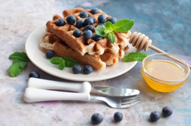 Beyaz tabakta yaban mersinli Belçika waffle 'ı, bal ve nane şekeri. Tatlı waffle 'lı kahvaltı. Beyaz saplı çatal ve bıçak.