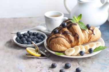 Çikolatalı kruvasan, yaban mersini, muz ve açık gri arka planda naneli. Kruvasanla kahvaltı, beyaz çaydanlık ve limonlu çay.. 