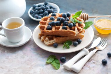 Beyaz tabakta yaban mersinli Belçika waffle 'ı, bal ve nane şekeri. Beyaz çaydanlık ve tatlı waffle ile bir fincan çayla kahvaltı. Beyaz saplı çatal ve bıçak.
