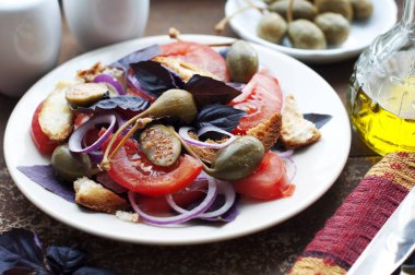 Ekmek, domates, fesleğen, kapari, kırmızı soğan ve koyu kahverengi arka planda beyaz tabakta zeytinyağı ile İtalyan Panzanella salatası. Bir tabak geleneksel İtalyan salatası ve tuzluk.