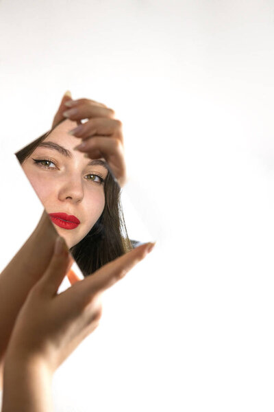 Концептуальный портрет молодой женщины с красной помадой, смотрящей в зеркало на белом фоне. Образ символизирует саморефлексию, идентичность, внутреннюю красоту, эмоциональную уязвимость и современный минимализм.