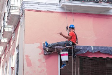 Miğfer ve emniyet kemeri takılı halat erişimi boyacısı, askıya alındığında binanın ön cephesine taze boya uygular, profesyonel bakım çalışmalarını yükseklikte gösterir