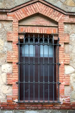 Çelik parmaklıklar ve dekoratif metal taçla birlikte tarihi taş pencereler, duvarcılıkta güvenlik sınırlamasını ve ortaçağ mimari mirasını ifade ediyor.