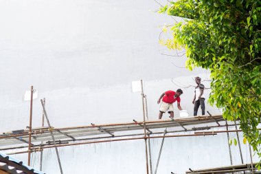 Maldivler, Thoddoo - 24 Şubat 2022: Gündelik iş kıyafetleri içinde koyu tenli ve şapkalı üç erkek işçi, inşaat işiyle meşgul, biri fırçayla ayakta duruyor, diğerleri beton ve ahşap panelli yapılara karşı iskelede oturuyor veya yaslanıyor