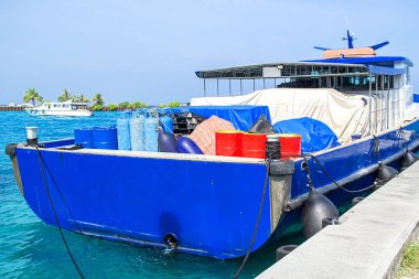 Mavi dalış botu, turkuaz denizdeki beton rıhtıma demirlemiş. Tüplü tanklar, yakıt varilleri ve kapalı teçhizatla. Uzak dalgalar ve açık mavi gökyüzünün altında palmiye ağaçları. Dalış gezileri, seyahat turizmi ve refah için tropikal ada maceralarını yakalar
