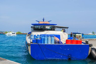 Maldivler, Dhigurah - 15 Şubat 2022: Kapalı benzin tanklarıyla dolu iskelede demirlenmiş mavi gemi, turkuaz su ve uzak ufukta karayı yöneten yelekli iki adam, adanın önemli ikmal operasyonlarını mercan bölgesinde resmediyorlar. Yüksek kalite fotoğraf.