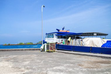 Maldivler, Dhigurah - 15 Şubat 2022: Mavi gemi limana demirlemiş, branda kaplı bölge, mavi gaz tankları, kırmızı variller ve doldurulmuş sandıklar yükleme veya boşaltmayı bekleyen portakallarla dolu, güvertede insan, turkuaz lagünü ve uzak ufukta