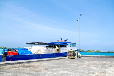 Mavi kargo teknesi turkuaz denizdeki beton rıhtıma demirlemiş kırmızı ve mavi varillerle dolu, yükleme için sandıklar dolusu portakal ve kapalı erzak. Uzak ufuk ve açık mavi gökyüzü. Otantik tropikal ada ticaretini, hazır kasalarla yakalar.