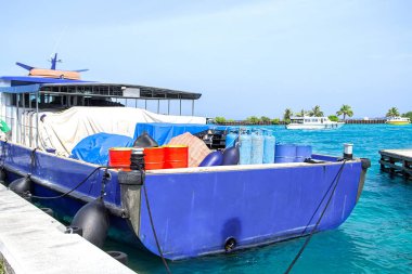 Maldivler, Dhigurah - 15 Şubat 2022: Kapalı benzin tanklarıyla dolu iskelede demirlenmiş mavi gemi, turkuaz su ve uzak ufuk yakınlarındaki kargoyu yöneten yelekli iki adam, mercan adasındaki önemli ada ikmal operasyonlarını resmediyorlar.