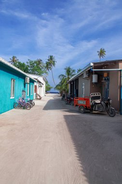 Maldivler, Thoddoo. Tropikal adanın dar kumlu sokaklarında renkli turkuazlar, bej binalar, palmiye ağaçları, park edilmiş bisikletler ve üç tekerlekli bisikletler var. Mavi gökyüzünün altında okyanus manzarası. Huzurlu ada yaşamını seyahat ve turizm için yakalar