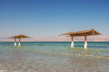 İsrail, Ein Bokek -26 Mar 2011: İki ahşap pavyon ve sazdan çatıları turkuaz Ölü Deniz 'in su bacakları İsrail sahillerinde beyaz tuz formasyonlarıyla kaplanmış Ürdün kıyılarına bakıyor. Seyahat için eşsiz doğal tuz fenomenini yakalar
