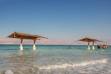 İsrail, Ein Bokek -26 Mar 2011: İki ahşap pavyon ve sazdan çatıları turkuaz Ölü Deniz 'in su bacakları İsrail sahillerinde beyaz tuz formasyonlarıyla kaplanmış Ürdün kıyılarına bakıyor. Seyahat için eşsiz doğal tuz fenomenini yakalar